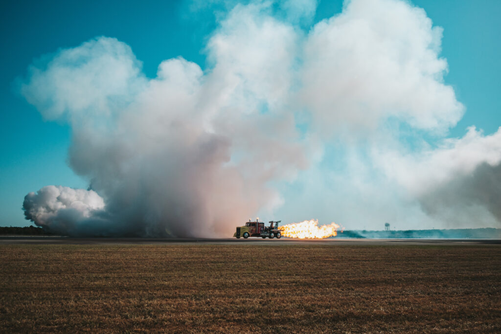 A truck with a mounted jet engine on a runway, shooting a massive orange and yellow flame horizontally across flat, brown earth. A teal sky and white clouds frame the scene.