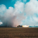 A truck with a mounted jet engine on a runway, shooting a massive orange and yellow flame horizontally across flat, brown earth. A teal sky and white clouds frame the scene.