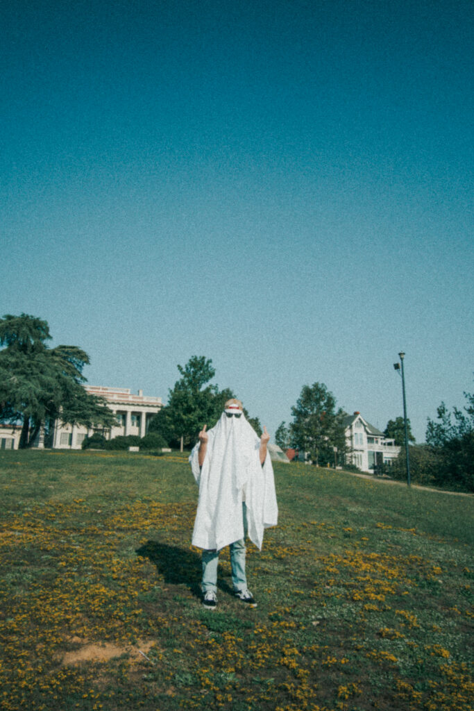 A figure wrapped in a white sheet stands in a field dotted with yellow wildflowers, with residential buildings visible in the background under a clear teal sky.