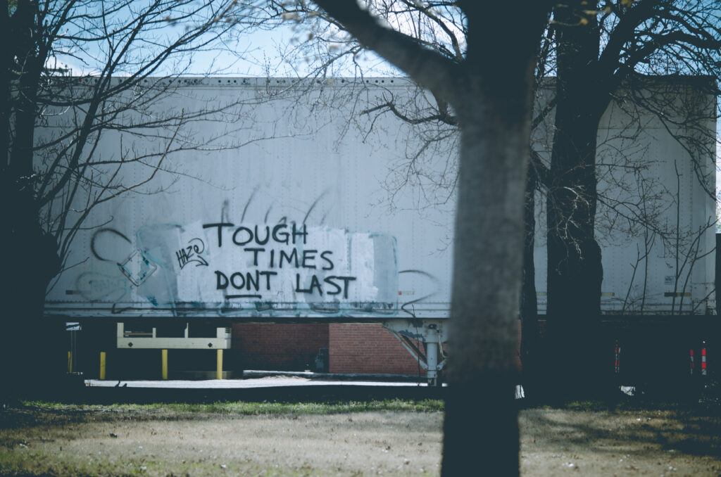 A photograph taken through bare tree branches of a box truck with graffiti reading "Tough Times Don't Last" in black spray paint on its white side. Industrial buildings and bare trees surround the scene.