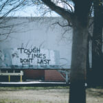 A photograph taken through bare tree branches of a box truck with graffiti reading "Tough Times Don't Last" in black spray paint on its white side. Industrial buildings and bare trees surround the scene.