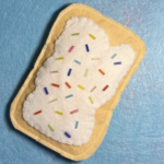 A handmade sewn soft sculpture shaped like a pop tart with a golden yellow felt crust and cream felt filling. The surface is decorated with scattered colorful glass seed beads in red, blue, yellow, green, and pink arranged like sprinkles.