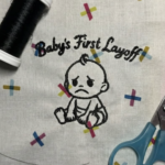 Machine-embroidered design on cream fabric showing a simple line-drawn baby with a sad expression, surrounded by colorful embroidered crosses in pink, yellow, and blue. Embroidery floss and scissors are visible at the edges of the composition.