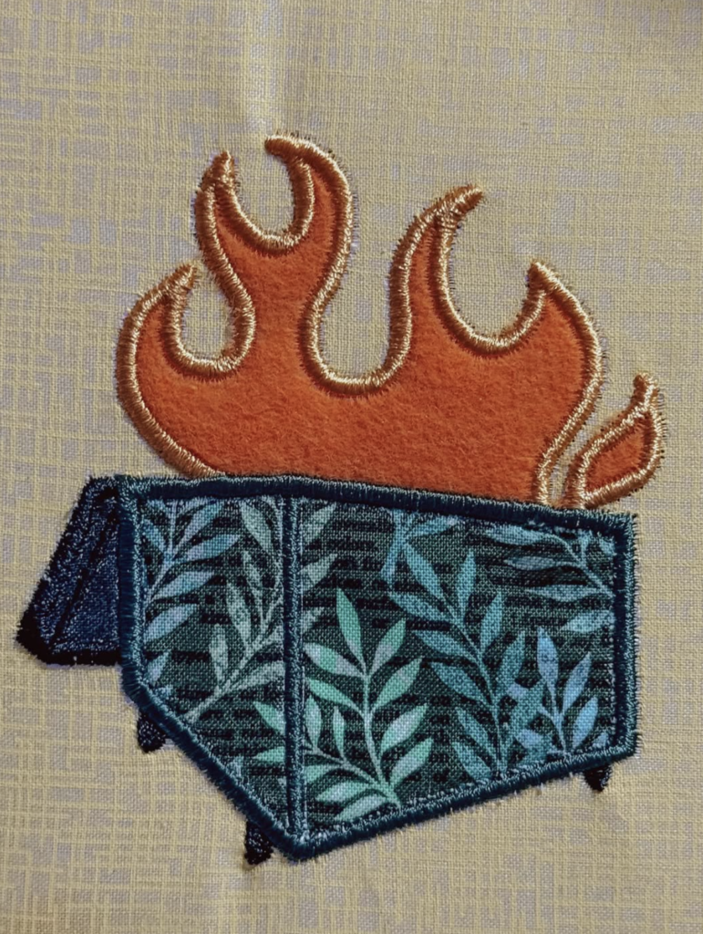 machine embroidered applique fabric piece featuring a rust and gold flame rising from a teal patterned garbage can against a yellow background. The flame has detailed embroidered edges and the pocket is filled with a leafy pattern in turquoise and navy.