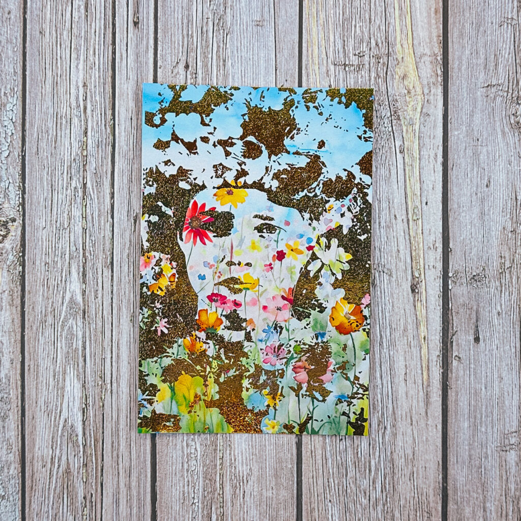 A vertical foil print resting on a light wood surface. The piece depicts the silhouette of a woman’s face and a massive, towering floral headdress. The background and hair are rendered in textured, shimmering gold foil, while the face and crown are filled with a colorful, watercolor-style floral pattern under a bright blue sky.