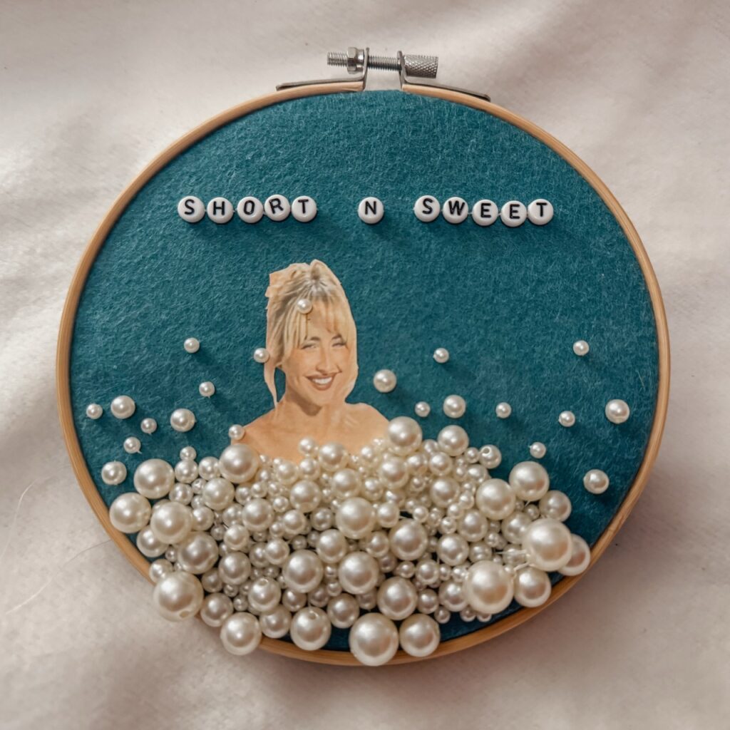 embroidery hoop on teal felt background featuring a portrait of sabrina carpenter embroidered with beads to replicate soap bubbles